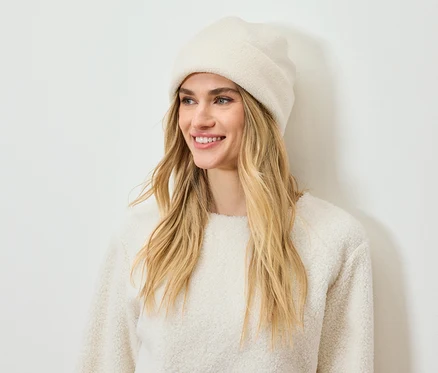 Une femme souriante aux cheveux blonds porte un bonnet et un pull crème.