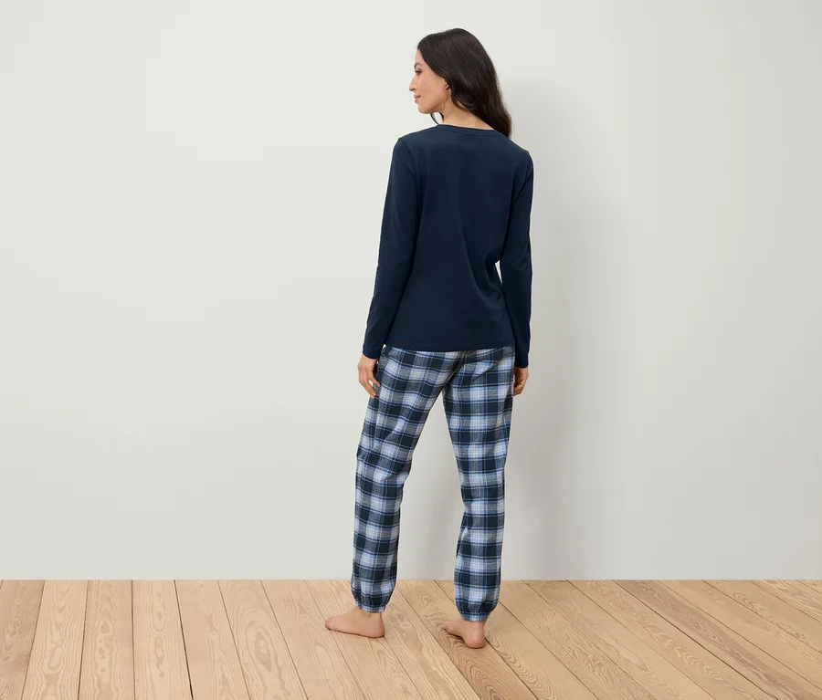 Une femme en pyjama bleu marine avec un pantalon à carreaux se tient dos à la caméra sur un plancher en bois devant un mur blanc.