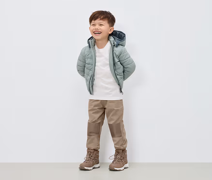 Un garçon souriant se tient debout, vêtu d'un manteau en duvet vert clair, d'un pantalon beige et de bottes marron.