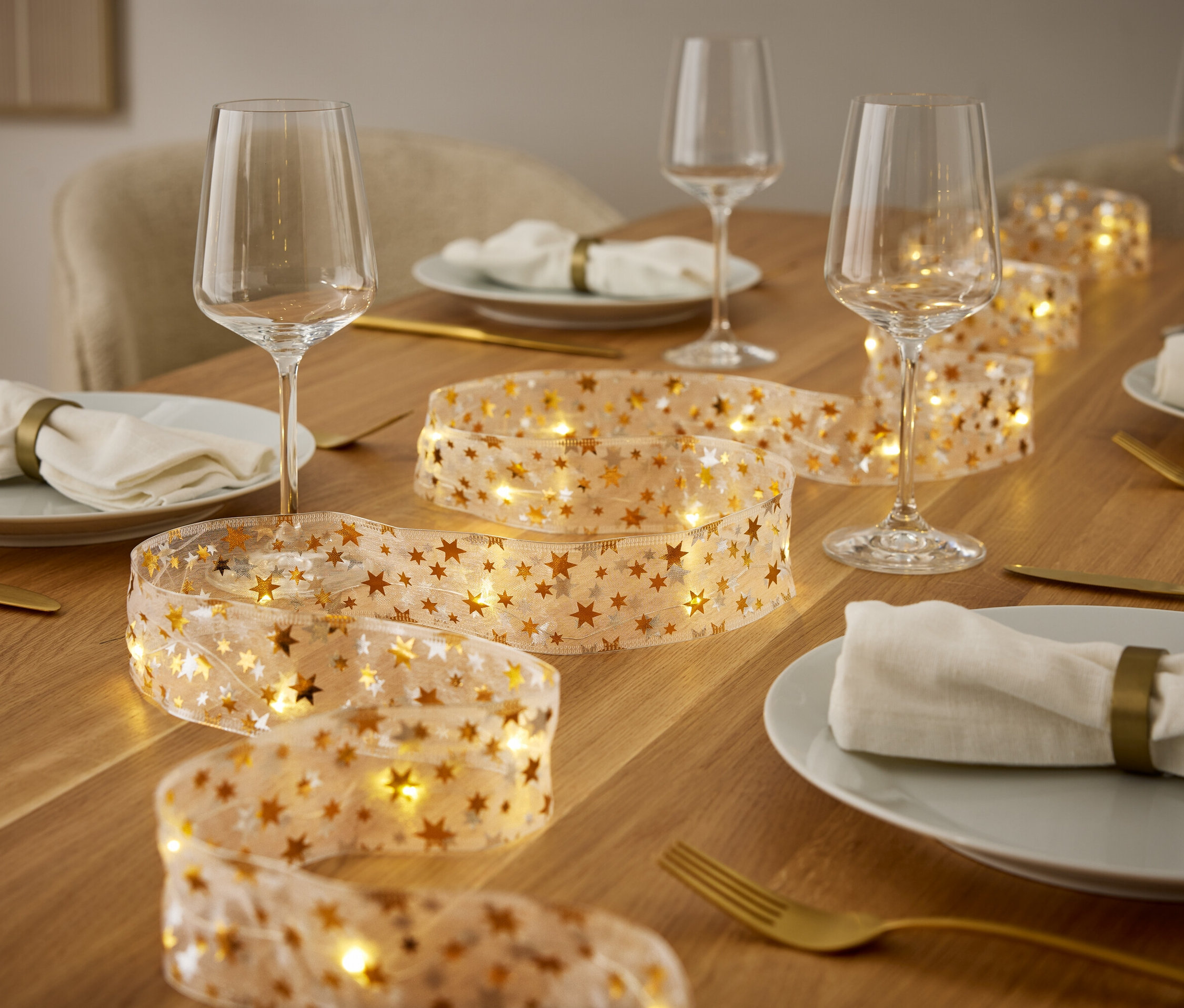 Une table dressée avec des assiettes, des verres à vin, des couverts dorés et une guirlande lumineuse avec des étoiles dorées.