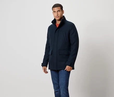 Un homme en manteau bleu marine et jean se tient devant un fond blanc.