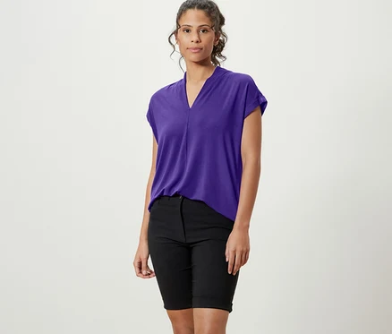 Une femme debout portant une blouse violette avec encolure en V, un short noir et des baskets blanches.
