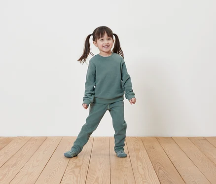 Une petite fille souriante avec des couettes pose dans un ensemble sweat-shirt et pantalon de jogging vert clair sur un plancher en bois devant un mur blanc.