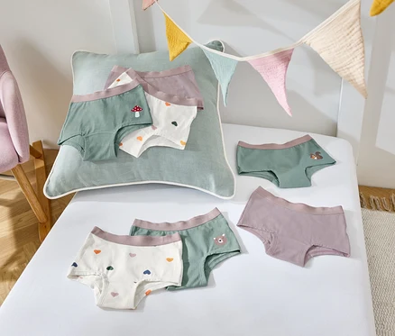 Sur un drap blanc sont disposés des sous-vêtements pour enfants de différentes couleurs et motifs, à côté se trouve un fauteuil violet.