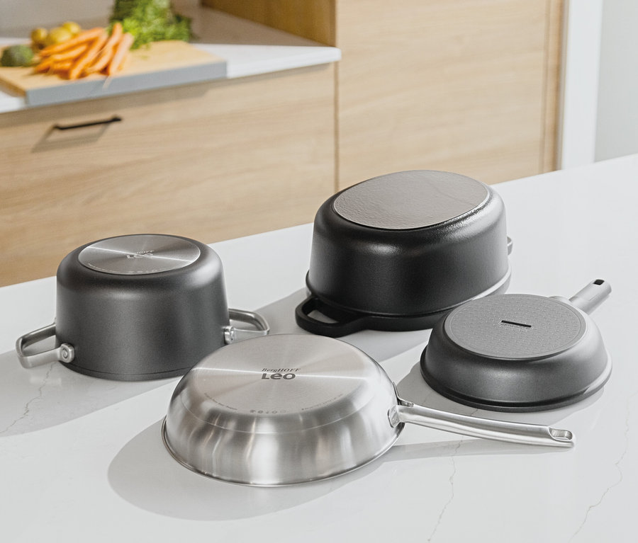 Quatre casseroles sont posées à l'envers sur un comptoir de cuisine blanc. À l'arrière-plan, on peut voir des armoires de cuisine en bois et des légumes sur une planche à découper.