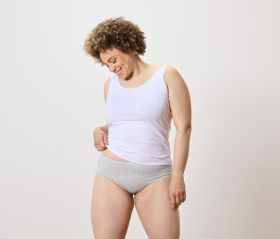 Une femme aux cheveux bouclés porte un débardeur blanc et une culotte à rayures grises et regarde vers le bas.