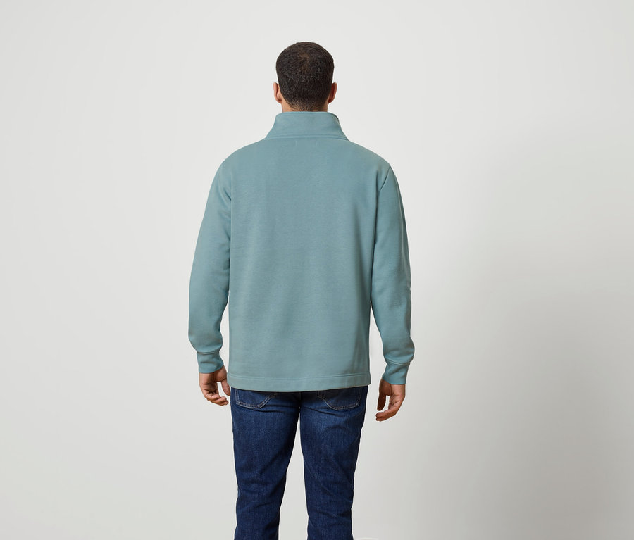 Un homme, vu de dos, porte un pull bleu-vert à fermeture éclair et un jean.