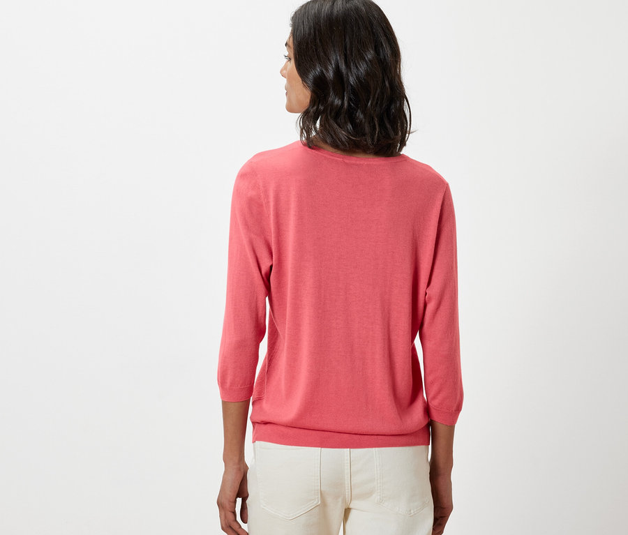 Une femme porte un pull-over corail en maille fine avec de la soie et un pantalon blanc.