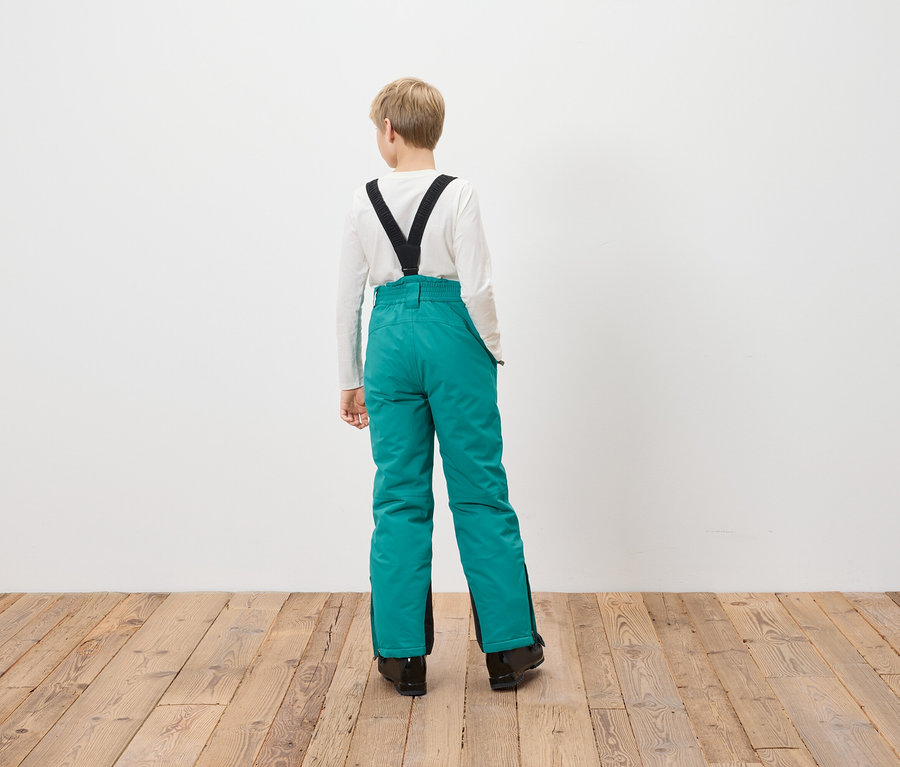 Un garçon se tient dos à la caméra portant un pantalon de ski turquoise avec des bretelles noires.