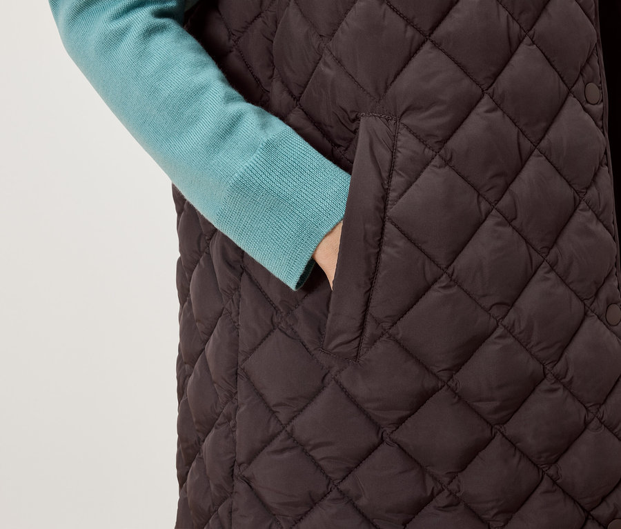 Main dans la poche d'un gilet matelassé marron, un pull bleu est visible sous le gilet.