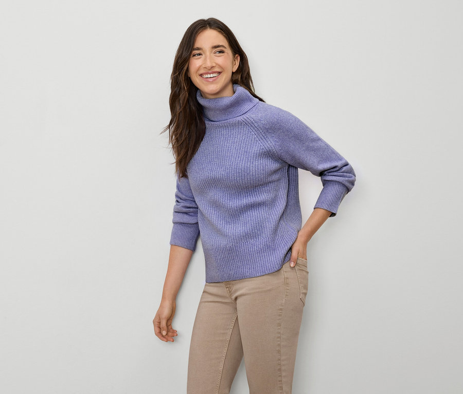 Femme souriante posant en pull à col roulé lilas et pantalon beige.