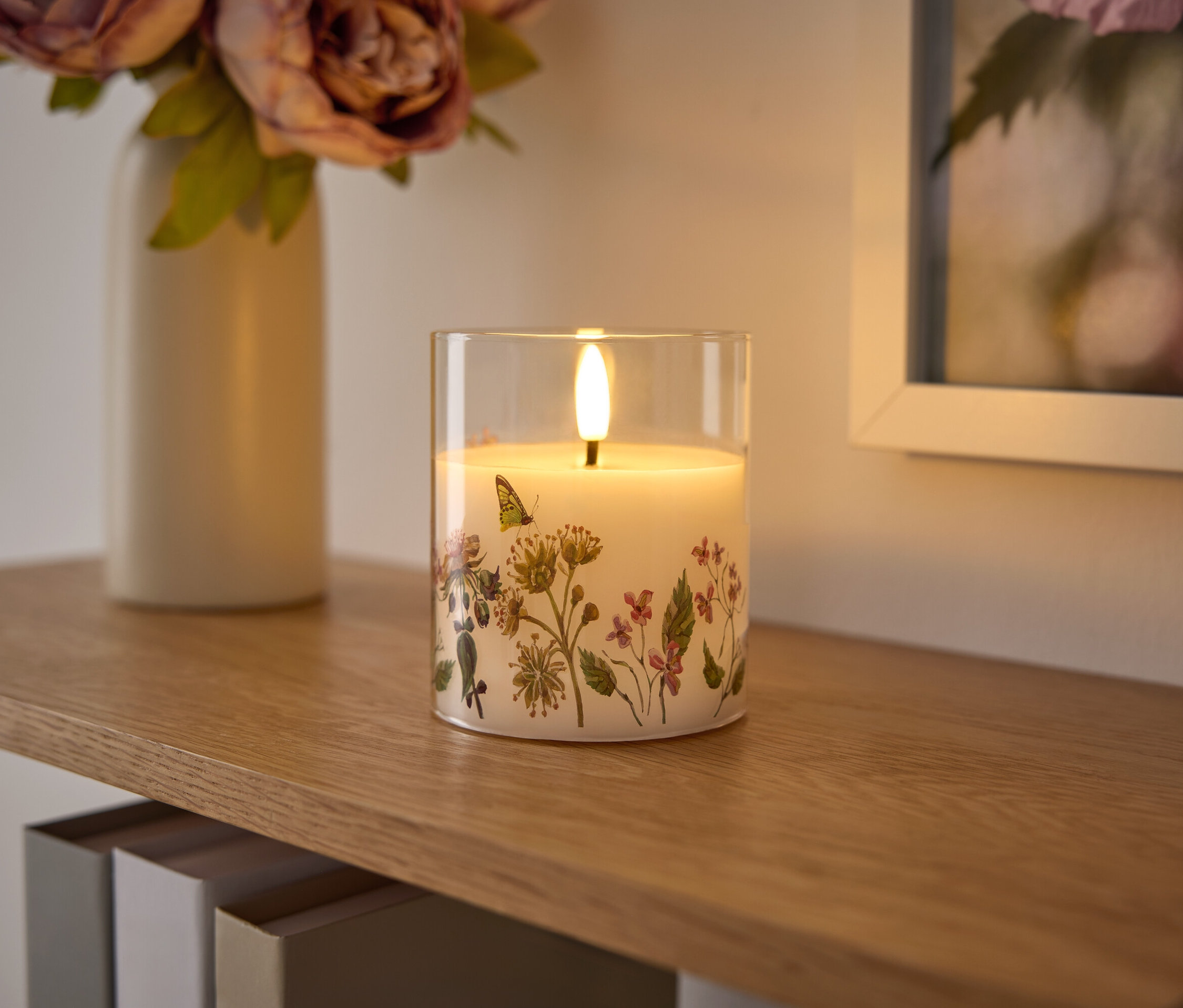 Sur une étagère en bois se trouvent une bougie en verre avec un motif floral, un vase avec des fleurs et une image encadrée.