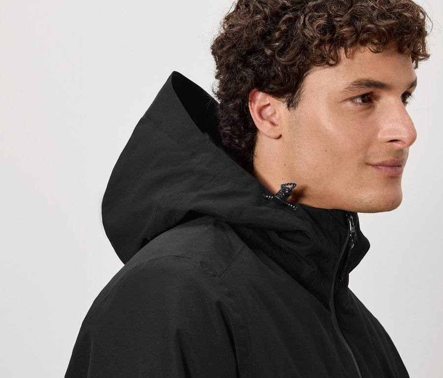 Un homme aux cheveux bruns bouclés porte une veste à capuche noire.