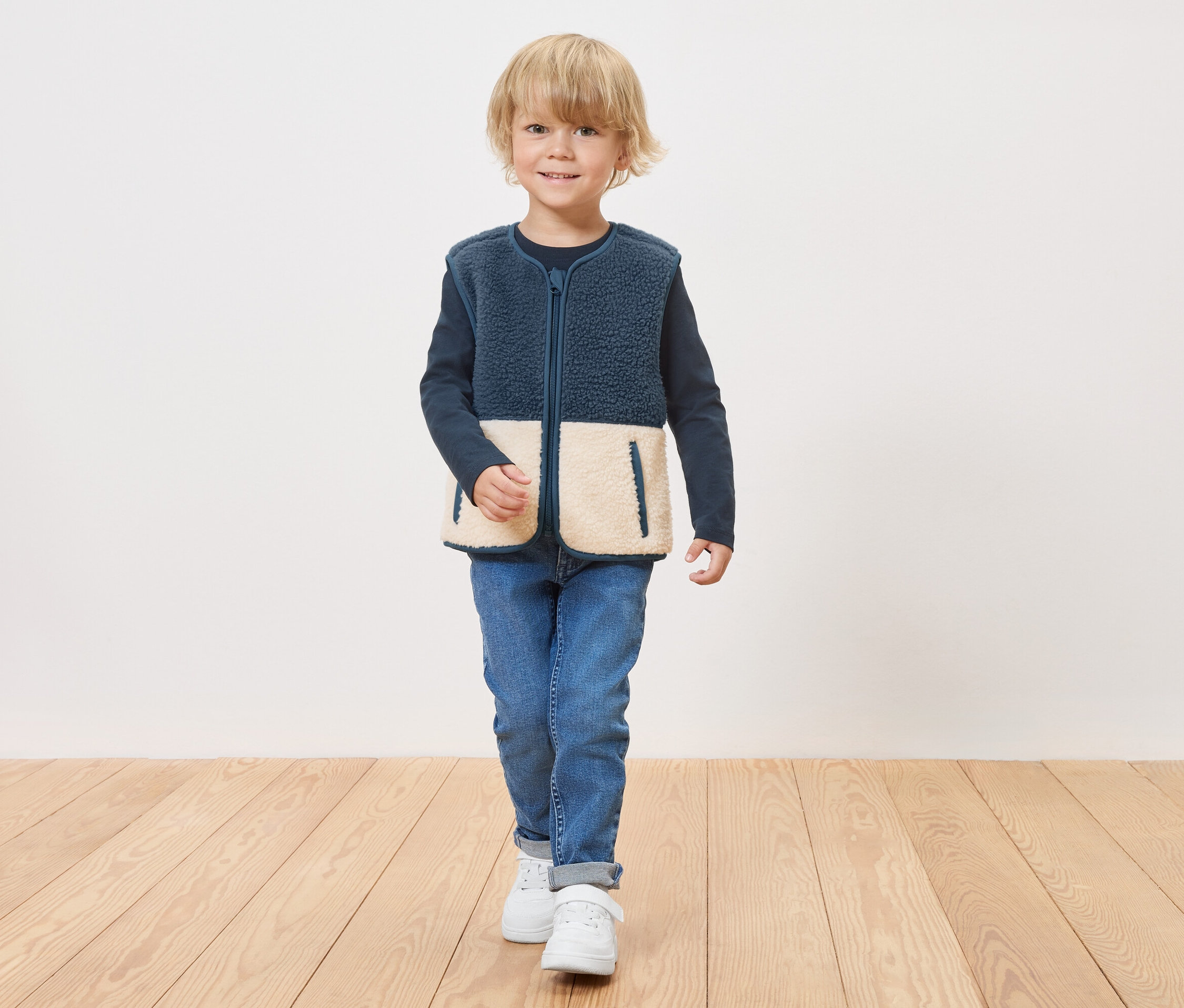 Un garçon portant un gilet en polaire bleu marine avec des empiècements inférieurs crème zippé se tient sur un plancher en bois.