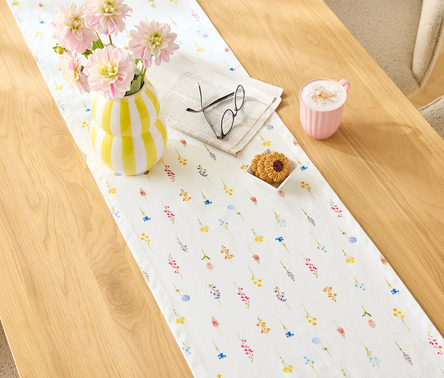 Sur une table en bois se trouve un chemin de table à motifs floraux, dessus un vase rayé jaune et blanc avec des fleurs roses, un journal avec des lunettes, des biscuits et une tasse rose.