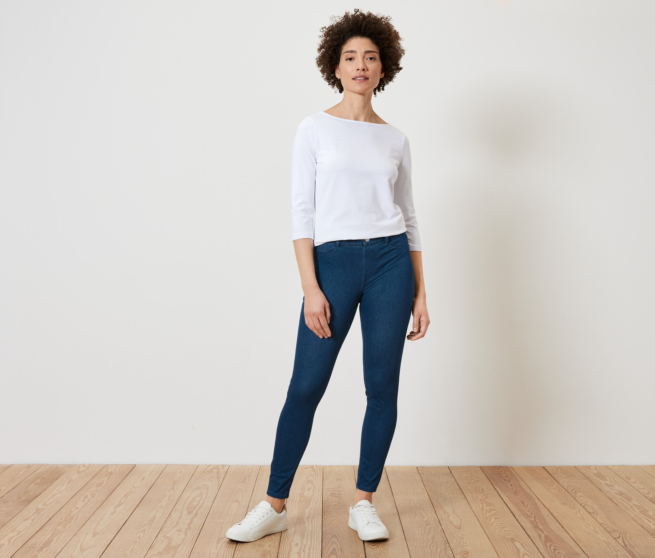 Une femme aux cheveux bouclés pose dans un haut clair à manches trois quarts, un jegging bleu foncé et des baskets blanches.