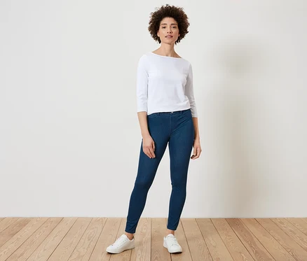 Une femme aux cheveux bouclés pose dans un haut clair à manches trois quarts, un jegging bleu foncé et des baskets blanches.