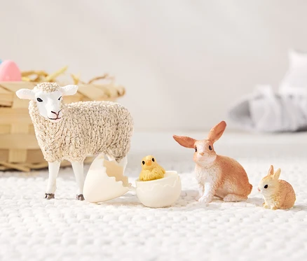 Un mouton se tient à côté d'une coquille d'œuf cassée d'où sort un poussin, à côté sont assis un lièvre et un petit lapin. À l'arrière-plan, il y a un panier en osier avec des bonbons et des baskets blanches. Set Schleich® «
Animaux de Pâques
».
