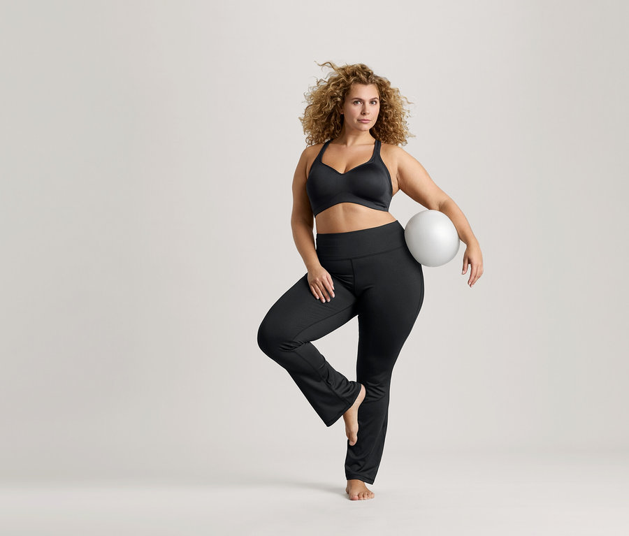 Femme aux cheveux bouclés en équilibre sur une jambe, tenant un ballon blanc, portant un soutien-gorge de sport noir et un legging noir.