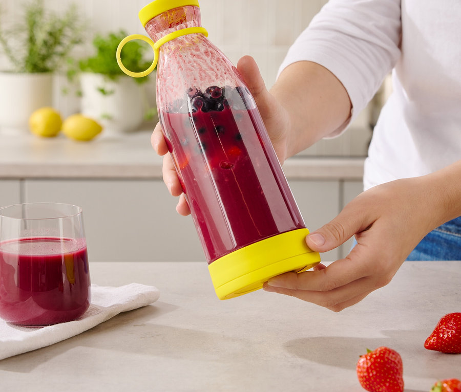 Une personne tient une bouteille transparente avec un couvercle et un fond jaunes, remplie d'une boisson rouge et de myrtilles.