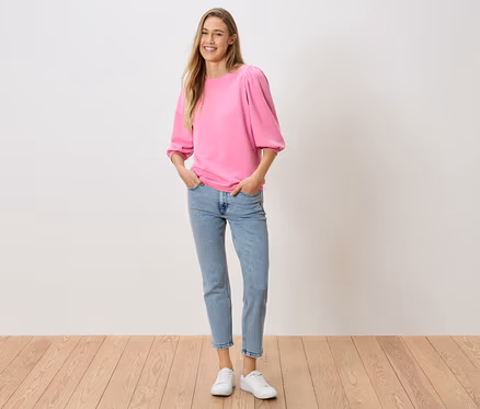 Une femme, les mains dans les poches de son pantalon, porte un haut rose à manches bouffantes, un jean et des baskets blanches.
