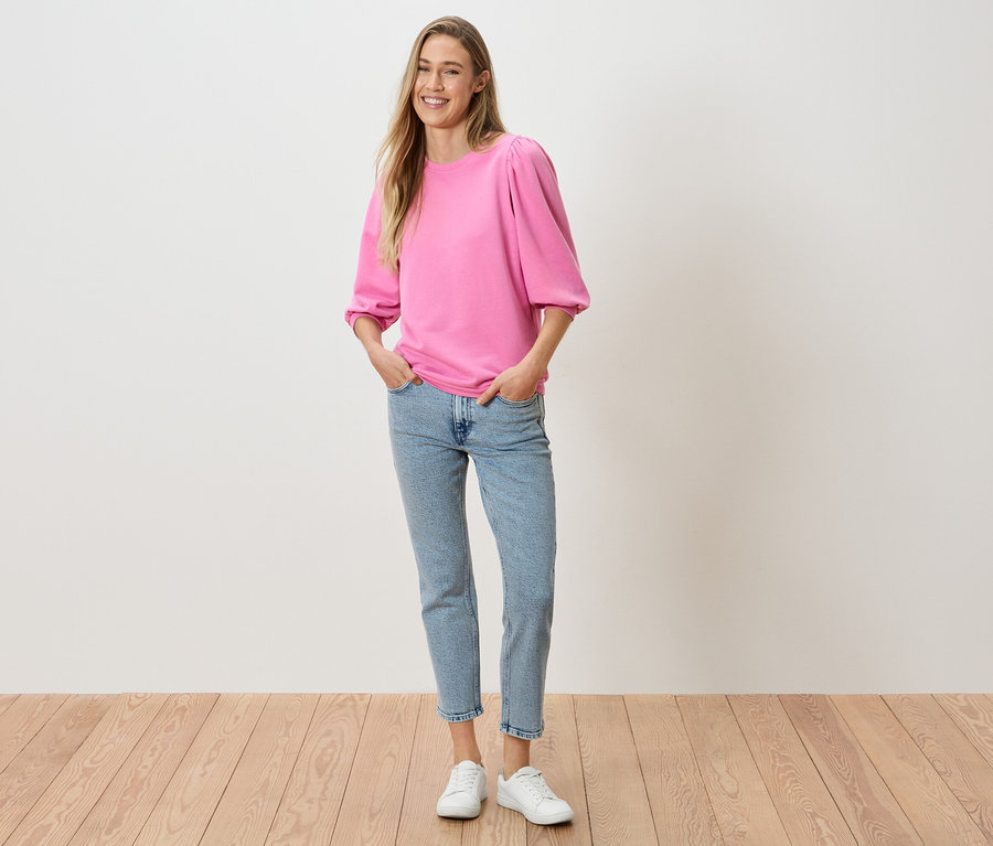 Une femme, les mains dans les poches de son pantalon, porte un haut rose à manches bouffantes, un jean et des baskets blanches.