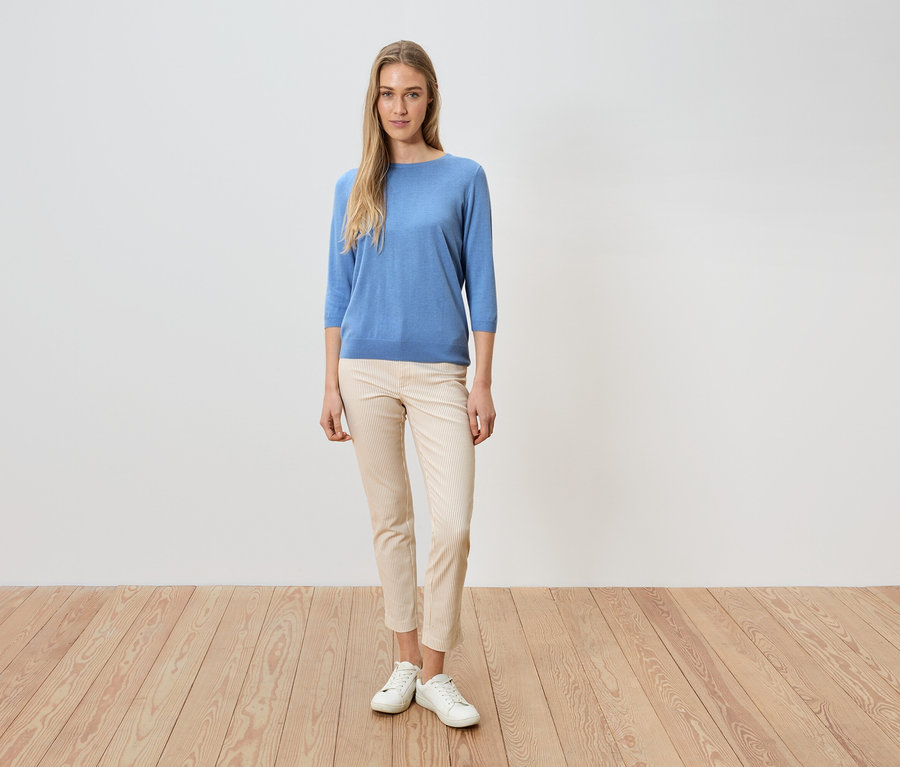 Une femme se tient devant un mur blanc, elle porte un pull bleu, un pantalon clair et des chaussures blanches.