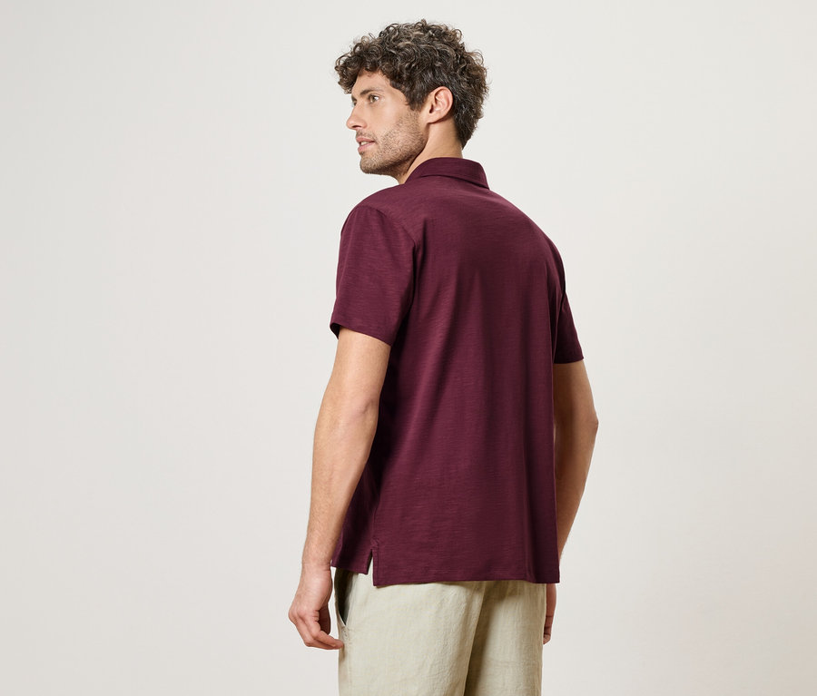 Un homme aux cheveux bouclés, vu de dos, porte un polo bordeaux et un short beige.