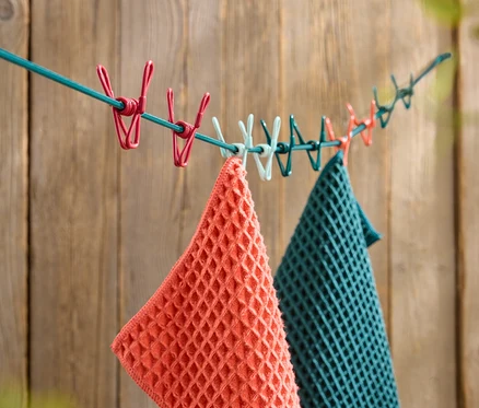 Une serviette gaufrée orange et une serviette gaufrée turquoise sont suspendues à une corde à linge avec des pinces à linge colorées devant un mur en bois.