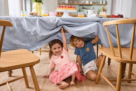 Deux enfants sont assis sous une table avec une nappe bleu clair. La fille porte un Kinder-Tüllrock et 2 Kinder-T-Shirts. Le garçon porte un Kinder-Shorts.