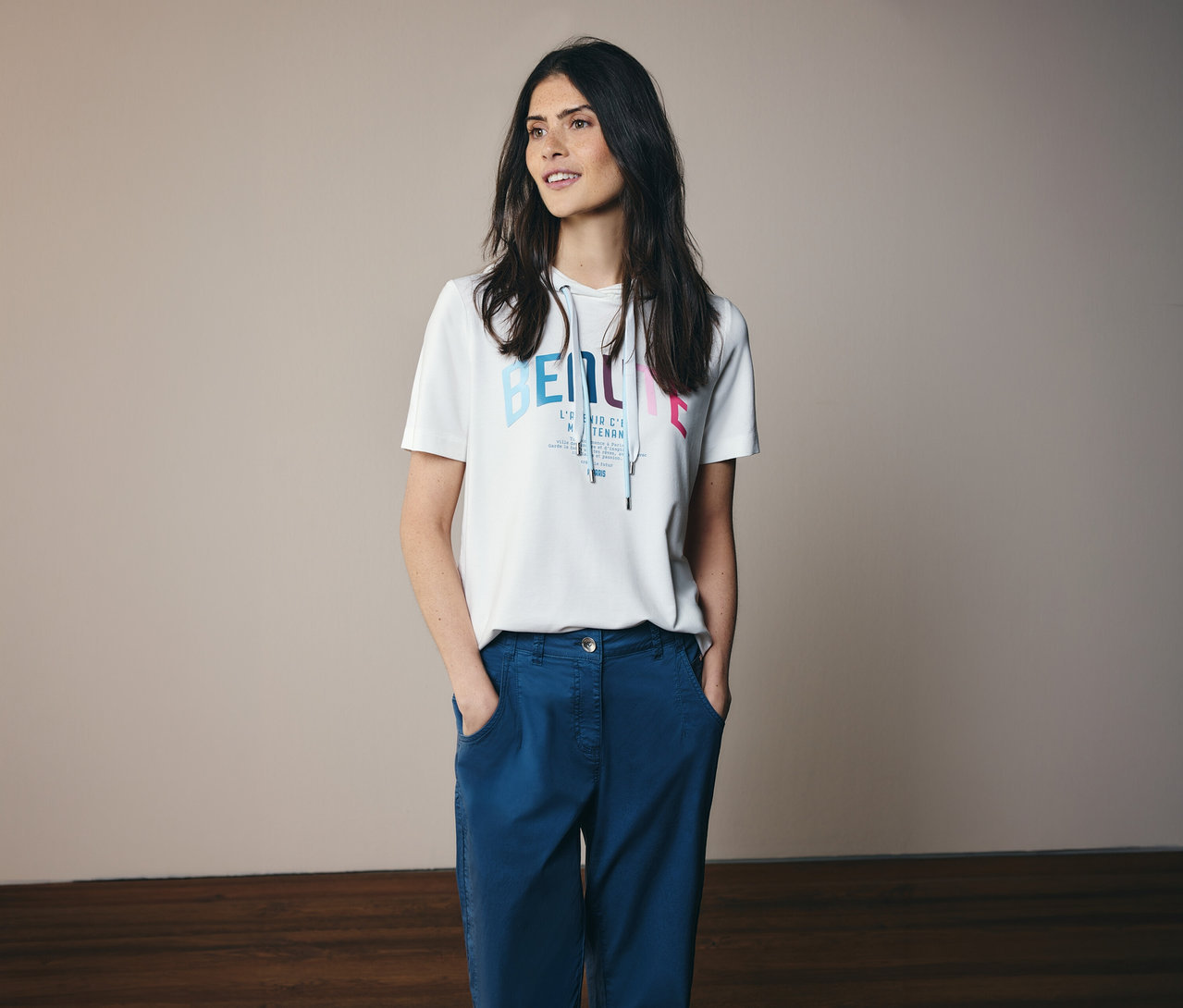 Femme portant un pantalon bleu et un t-shirt à capuche CECIL blanc avec un imprimé, les mains dans les poches.