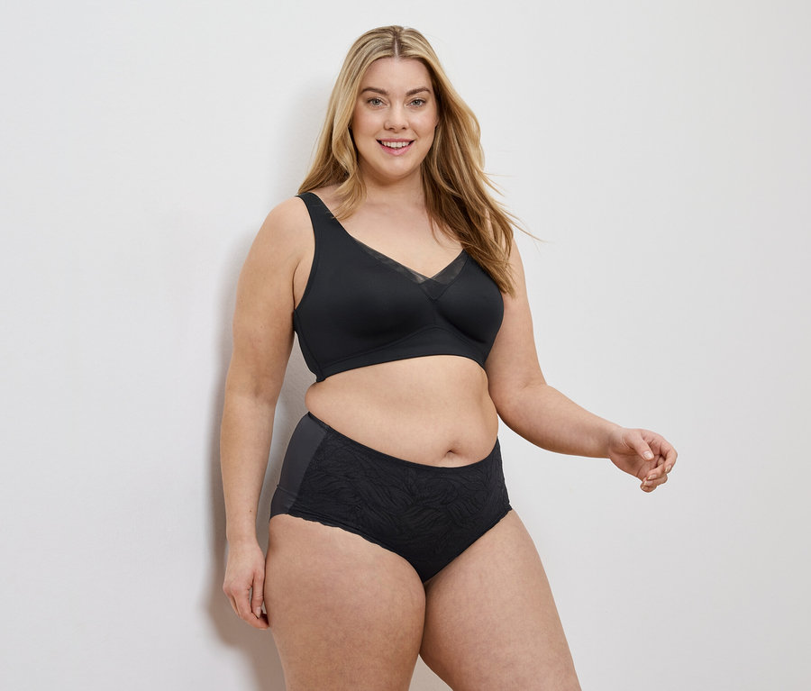 Une femme aux cheveux blonds mi-longs porte un soutien-gorge noir et une culotte noire sur un fond blanc.