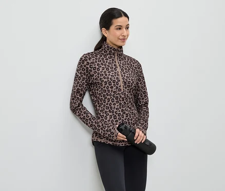 Femme portant un haut imprimé léopard marron, un legging noir et tenant une bouteille d'eau noire, appuyée contre un mur blanc.