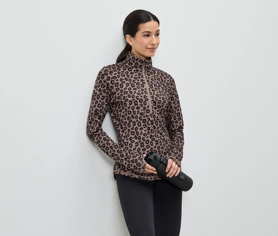 Femme portant un haut imprimé léopard marron, un legging noir et tenant une bouteille d'eau noire, appuyée contre un mur blanc.