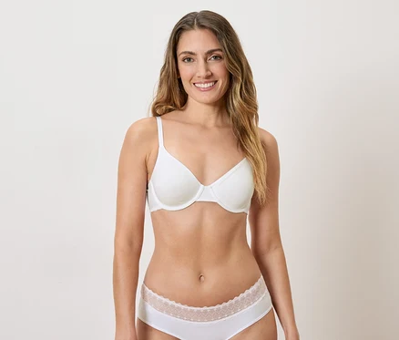 Une femme aux longs cheveux bruns ondulés portant un soutien-gorge blanc et des sous-vêtements blancs avec des détails en dentelle.