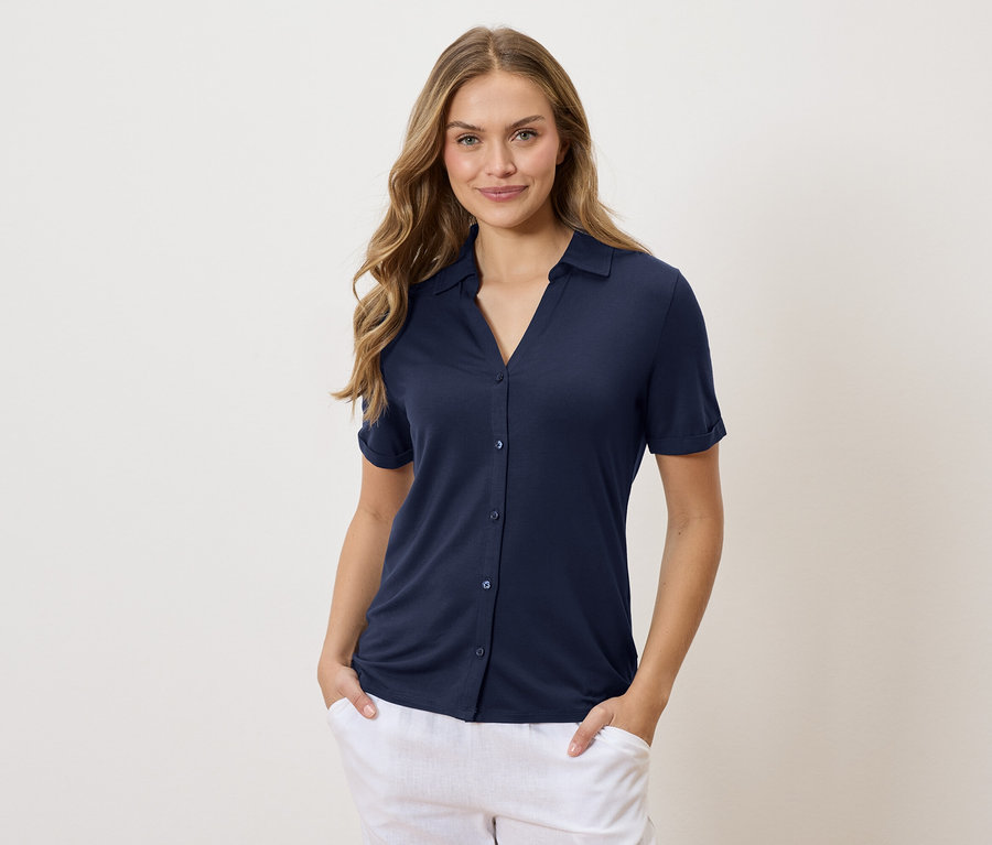 Femme aux longs cheveux blonds ondulés portant une chemise bleu marine et un pantalon blanc, les mains dans les poches.