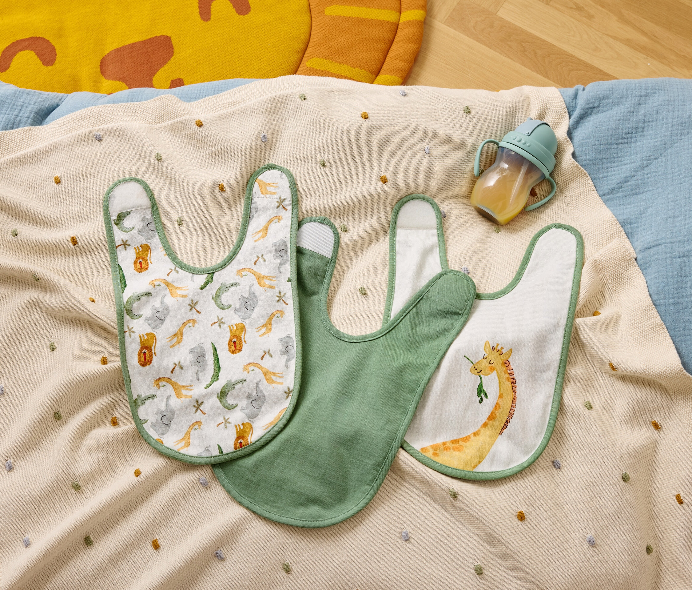 Trois bavoirs et un biberon sont posés sur une couverture beige à pois colorés.