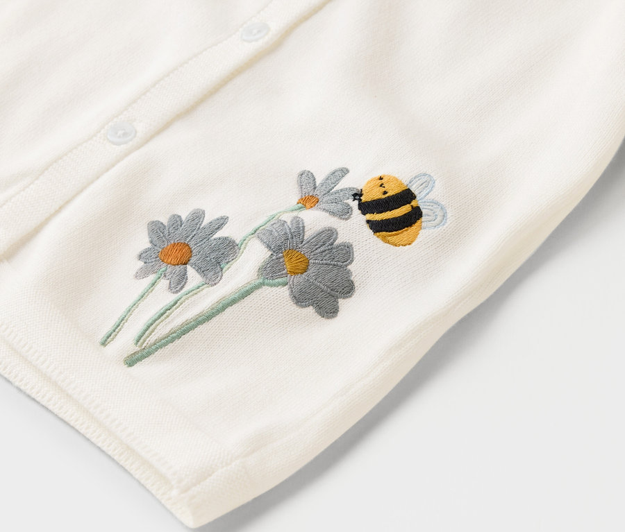 Gros plan sur un cardigan tricoté crème avec des boutons, brodé de marguerites et d'une abeille bourdonnante.