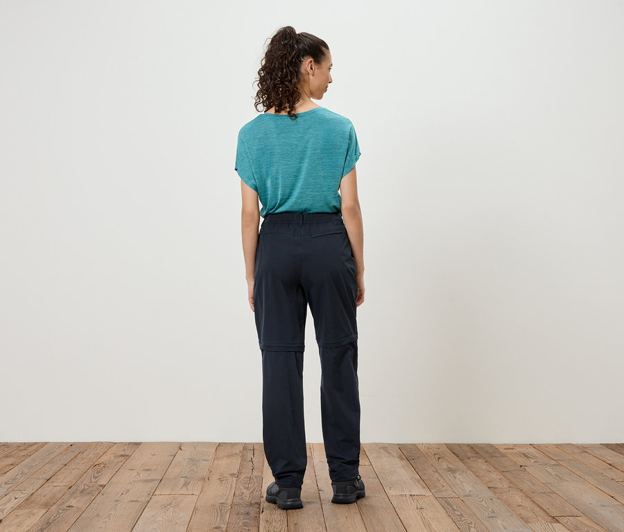 Femme debout dos à la caméra, portant un t-shirt bleu et un pantalon foncé avec des jambes zippées.
