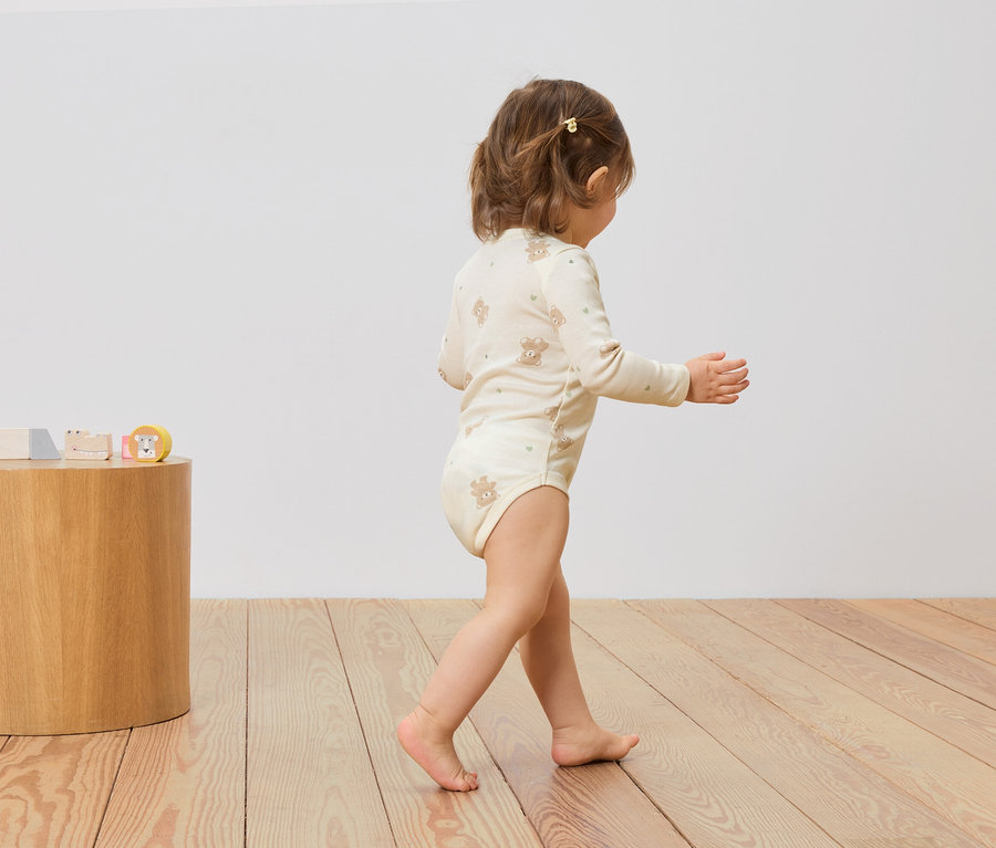 Un bébé en body beige à imprimé ours marche sur un plancher en bois. Des jouets en bois sont posés sur un tabouret en bois.