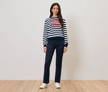 Une femme porte un pull rayé avec un imprimé, un jean foncé et des chaussures blanches.