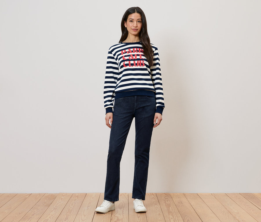 Une femme porte un pull rayé avec un imprimé, un jean foncé et des chaussures blanches.