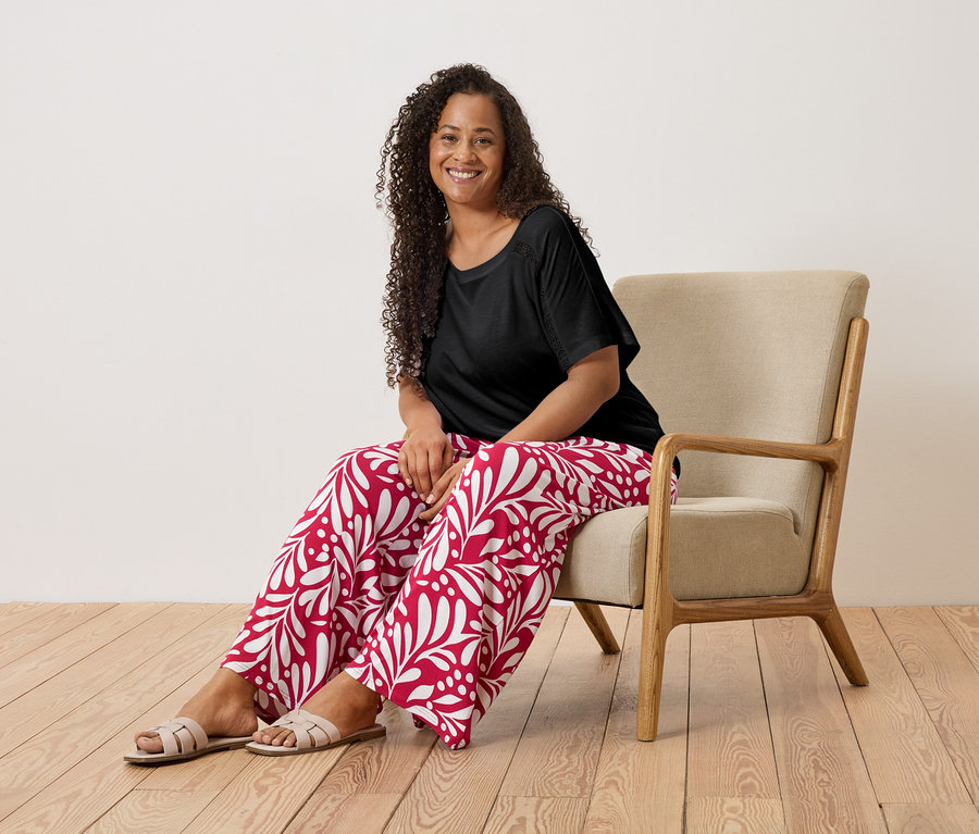 Une femme souriante aux cheveux bouclés est assise dans un fauteuil, portant un t-shirt noir et un pantalon rouge et blanc.