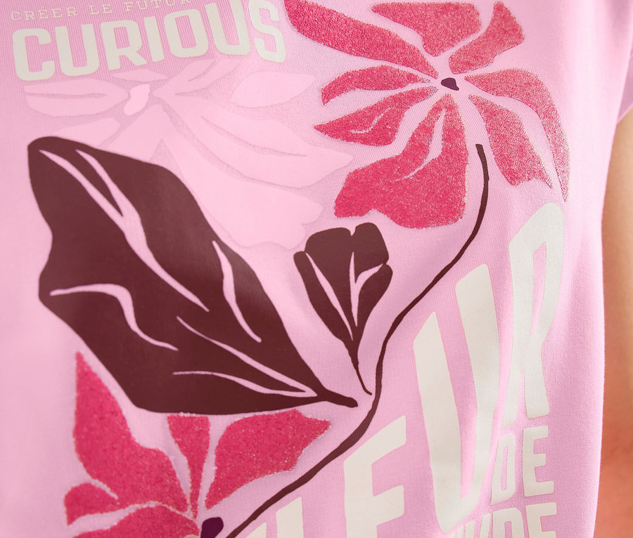 Une personne porte un T-shirt à motif CECIL rose avec un motif floral rose et marron foncé et les inscriptions "CURIOUS" et "FLEUR DE VIVRE".