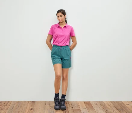 Une femme se tient devant un mur blanc, portant un polo rose et un short vert.