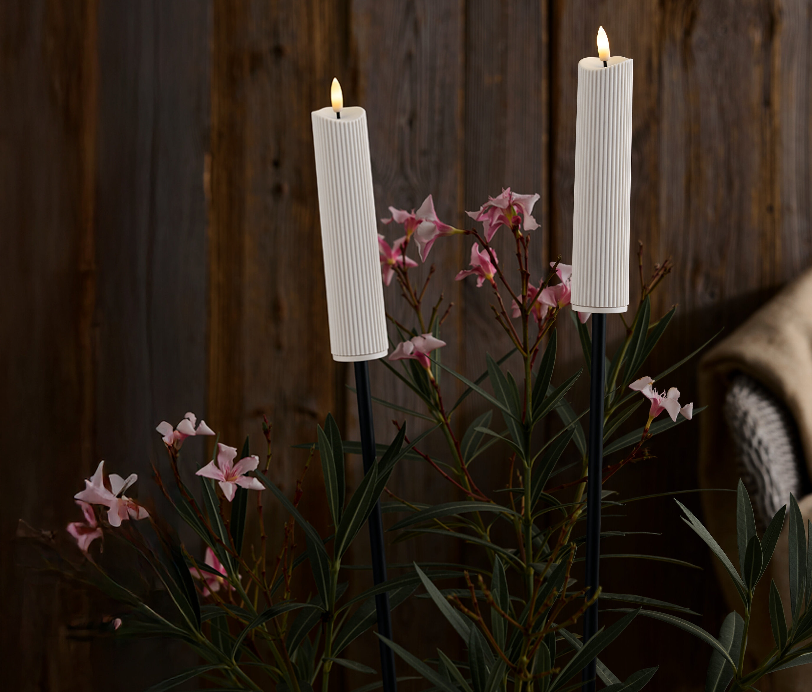 Deux bougies blanches côtelées sur des supports noirs au milieu de fleurs roses et de feuilles vertes devant un fond en bois foncé.