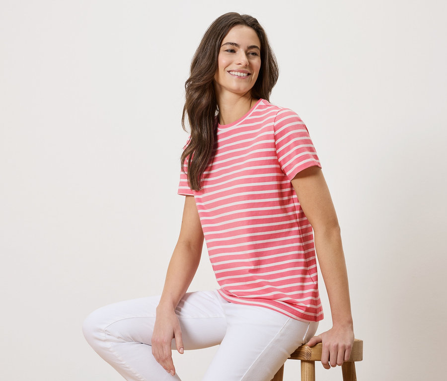 Une femme est assise sur un tabouret, portant un t-shirt rayé rose et blanc et un jean blanc.