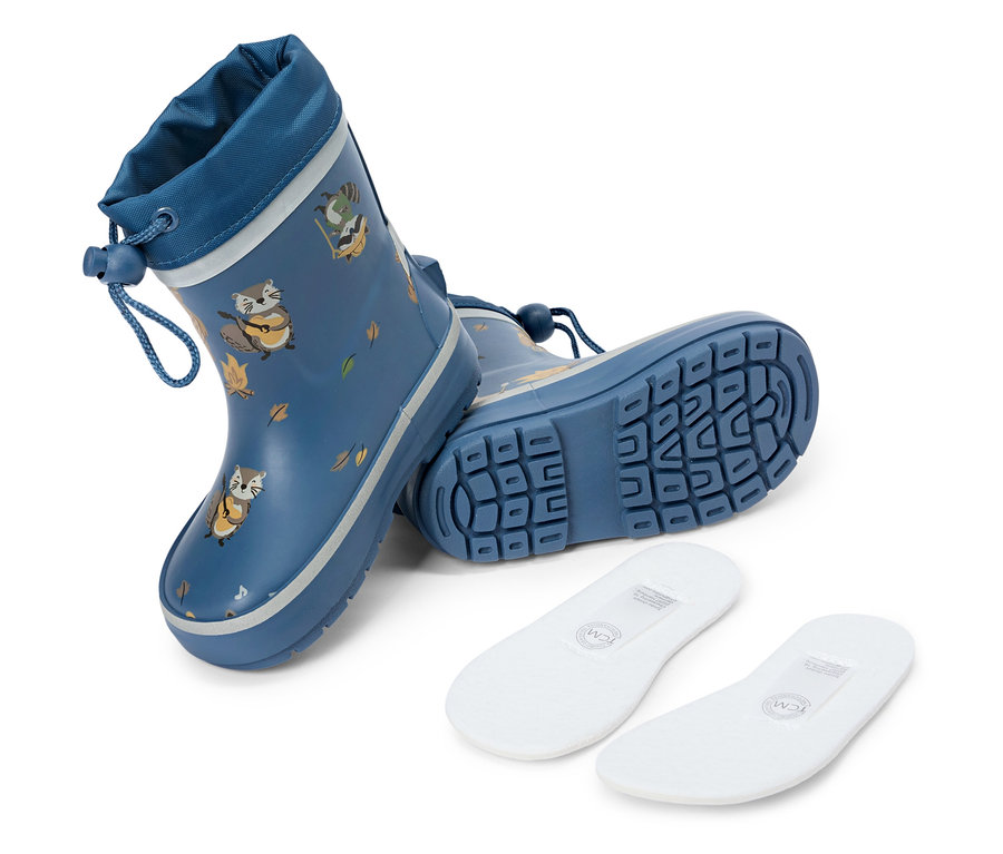 Une botte en caoutchouc bleue avec un motif de raton laveur est debout, une autre est couchée à côté. Deux semelles intérieures blanches sont au premier plan.