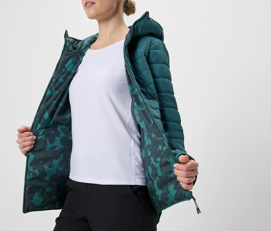 Femme portant une veste matelassée pétrole à capuche ouverte avec une doublure à motifs floraux sur un t-shirt blanc et un pantalon noir.