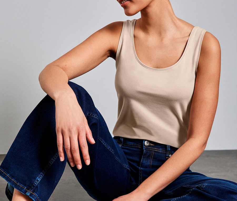 Femme aux cheveux courts et bouclés assise sur un sol gris, portant un Haut classique Street One beige et un jean large.